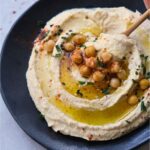 creamy hummus on a black plate with a drizzle of olive oil garnished with roasted chickpeas and parsley. Squeezing lime over the top.