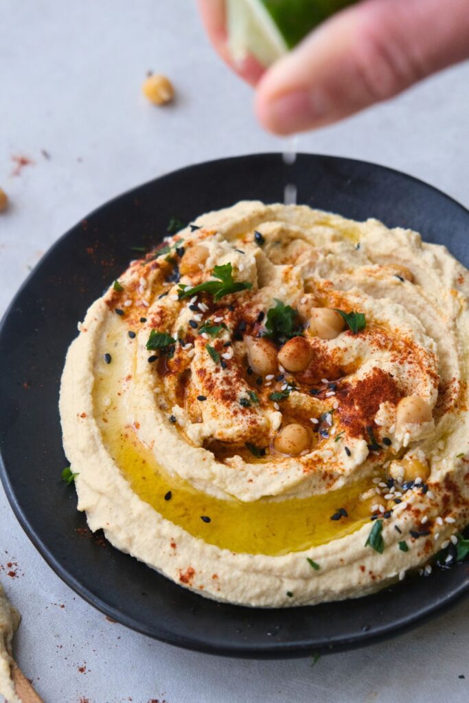 creamy hummus on a black plate with a drizzle of olive oil garnished with roasted chickpeas and parsley. Squeezing lime over the top.