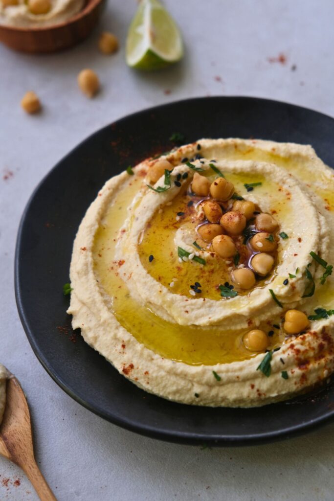 creamy hummus on a black plate with a drizzle of olive oil garnished with roasted chickpeas and parsley. Spoon with hummus in the corner.